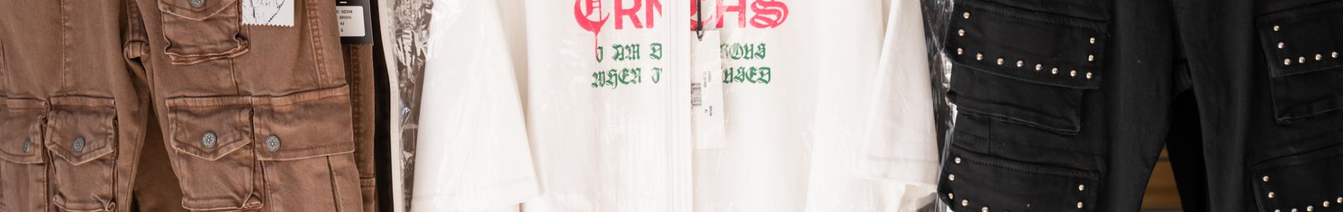 Display of clothing on hangers, featuring brown cargo pants, a graphic T-shirt in a plastic garment bag, and black pants.
