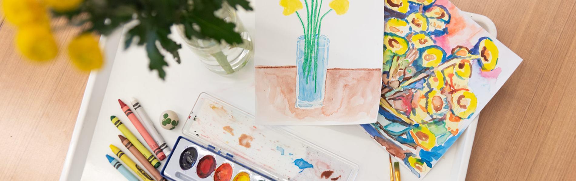 paints and painted items on a brown wooden table with a vase of yellow flowers